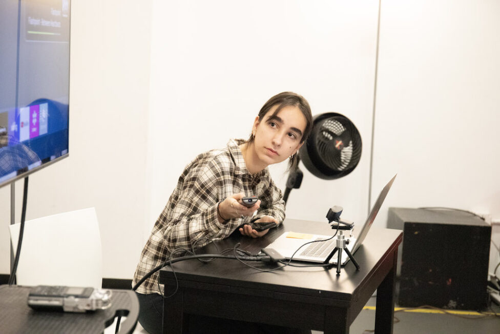Leonor leans over a desk with a remote in her hand. She sits in front of a laptop in a plaid top photo by Abigail Montes