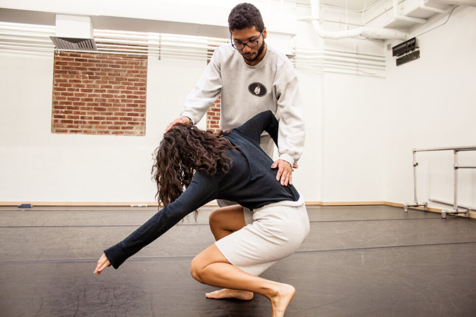 A photo from Rebecca Brooks' Alexander Technique Class. One dancers crouches while another places their hands on the first's head and lower back guiding them. Photo by Scott Shaw.