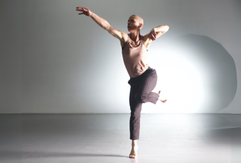 An action shot of Jennifer Nugent in a bright space. A light spotlights behind her as she holds her arm out and balances on one foot. She wears a pink top and grey striped pants. Photo by Jim Coleman.