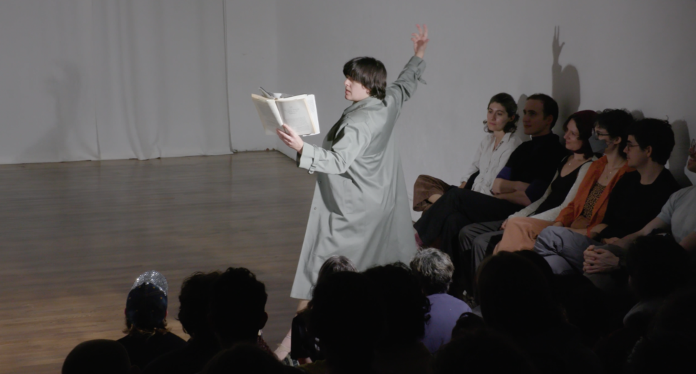 Tess Dworman fixes her gaze on the spine of a book as she holds it spread open with one hand and throws the other back triumphantly. Image by Kayhl Cooper. 
