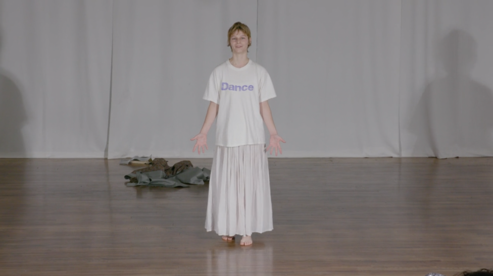 Sonya Gadet-Molansky stands barefoot with open palms in a moment during Everything Must Go. They wear a white t-shirt with 