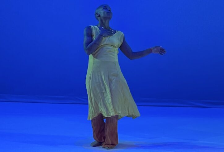 A blue lit stage with Jasmine Hearn in a yellow dress and orange pants. They look up with their hand to their chest one arm bent and out. Photo courtesy of artist.