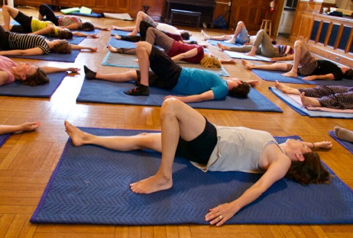 A photo of students practicing movement during a class. They lie on their backs on blue mats. One leg extended downward and the other leg was propped up with their knee bent and the foot pressed into the floor. Photo by Eve Boltax