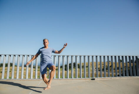 A brown-skinned Filipino man, Gerald wears a blue t-shirt and shorts and dances in front of a blue sky and grey fence. Photo by Crystal Birns