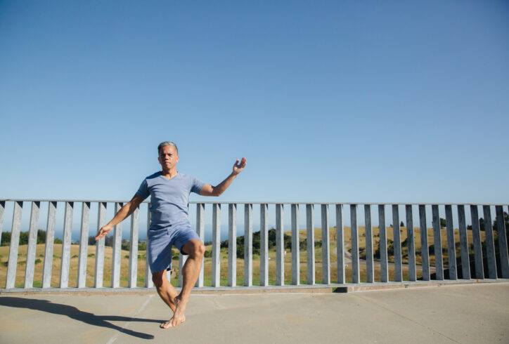 A brown-skinned Filipino man, Gerald wears a blue t-shirt and shorts and dances in front of a blue sky and grey fence. Photo by Crystal Birns