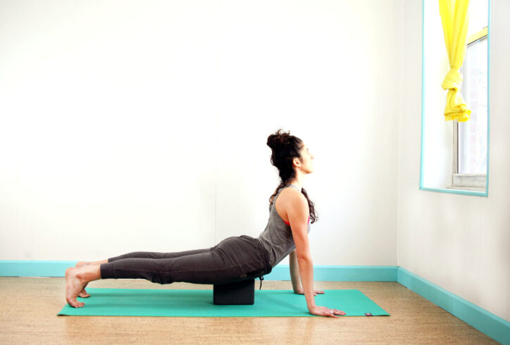 Light skin female is in a plank like position with a block supporting her pubic bone for support. Her chest is extended out and up to a window. Kay Ottinger. Photo Courtesy of artist.