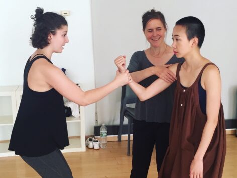 Emily stands between two female students, one who is making a fist, and the other who is holding the forearm of the one making the fist. The student who is grasping the forearm expresses surprise and the other calm. Emily is smiling, and touching the arm of the student making the fist. Photo courtesy of the artist.