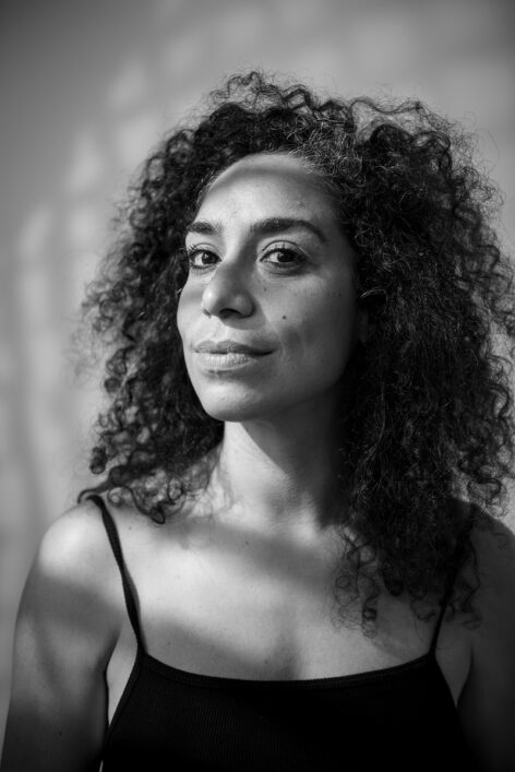 Headshot of Marina Santo. Shadows fall over her face as she smiles softly. She has voluminous curly hair. Photo by Fernanda Carvalho.