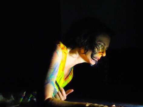 Nicole, a white woman with curly black hair and hazel eyes is prone, propped on her forearm with a pool of white light shining on her face. The light glows on her neck, a colorful abstract arm tattoo, and her fluorescent yellow dress. Photo by Brad Burkons.