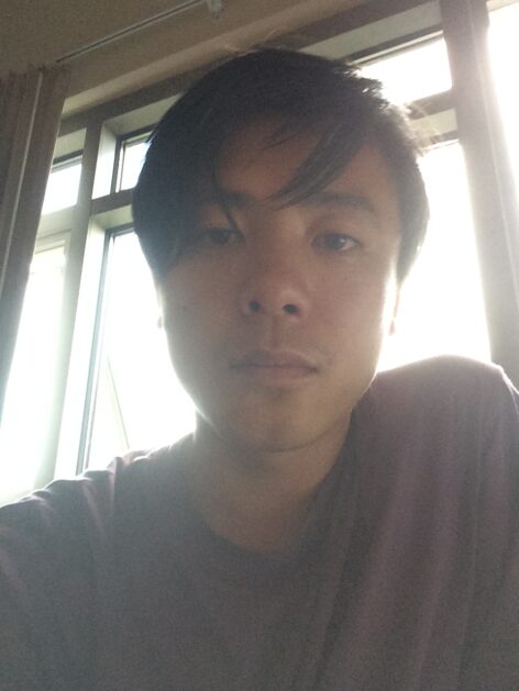 Jian Yi, an East Asian person looks at the camera with short length black hair and purple T-shirt in a portrait profile shot as light shines through a window behind them. Photo courtesy of the artist.
