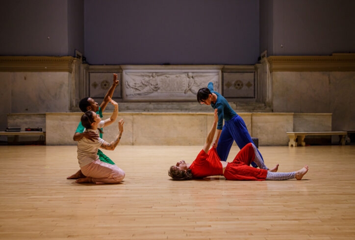 A photo from Jade Manns' performance for Movement Research at the Judson Church; two dancers kneel and gesture with their arms reaching upward and their palms facing out; two other dancers are engaged in a movement; one dancer wearing red lies on the floor and reaches towards the second dancer wearing blue, and the second dancer leans over pulling the first dancers shirt. Photo by Rachel Keane
