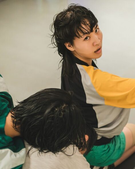 An intimate portrait of a person with wet, tousled black hair styled in a mullet, turning to face the camera with a direct, expressive gaze. They wear a sporty outfit—black, yellow, white, and gray top with green shorts—conveying a casual, athletic vibe. The softly blurred background draws attention to the subject’s presence, emphasizing the emotional intensity and texture of the moment. The image feels raw and editorial, capturing a sense of vulnerability, strength, and quiet intensity. Photo by @friezeofficial
