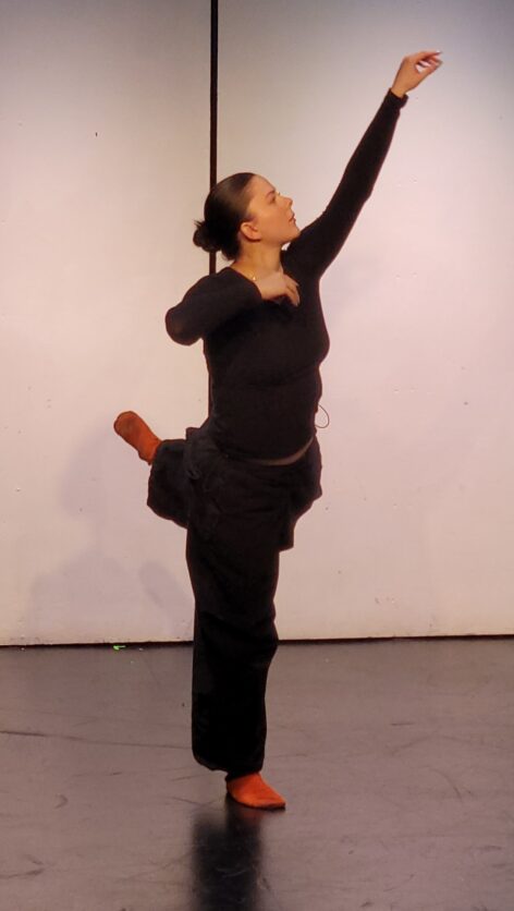 Rachel balances on one leg, hovering the other leg behind her in attitude. She wears all black with bright orange socks. She reaches over head, extending her arm softly above her. Photo by Maura Nguyen Donohue.