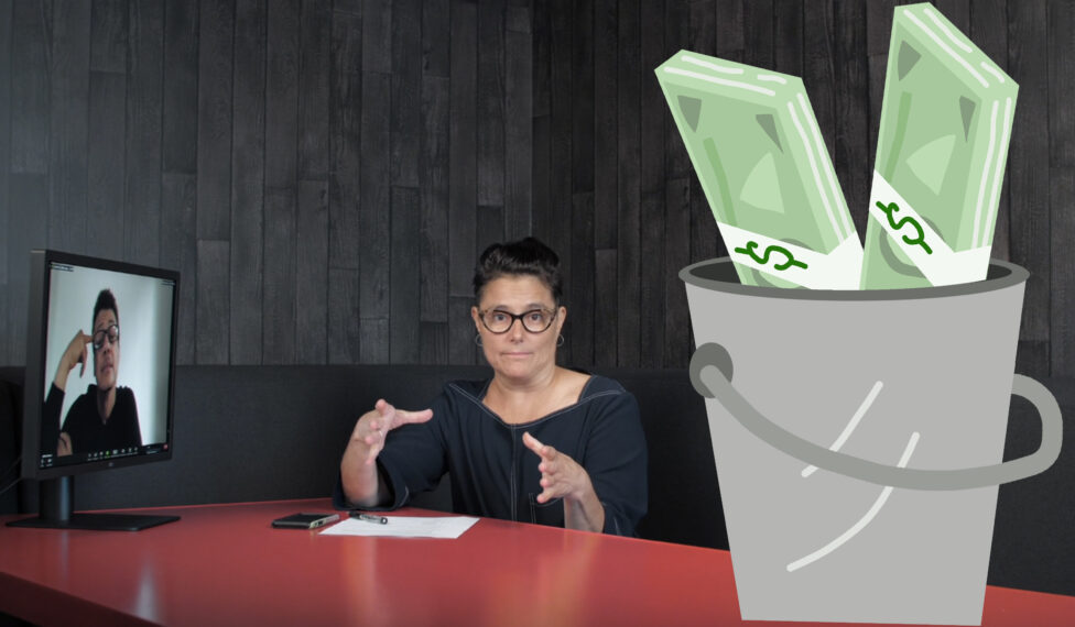 A screen capture of a video of a woman at a desk seated at a desk. Another person in a virtual meeting on the screen. They both wears glasses and a black top. A cartoon image on a grey bucket with money sit on the desk. Photo provided by Artists.