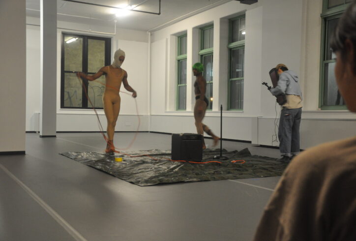 Three performers set up on a square tarp, they have cords and a large amp in the center of them. Artists are unknown. Photo by Mary Anne Bodnar.