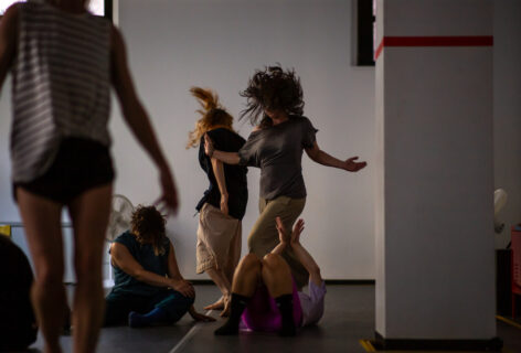 During a workshop in the Courtyard Studio. luciana teaching with hair flying upwards hiding face and arms and hands open to sides. Some students on the floor with arms up, others upright in movement. Photo by Rachel Keane