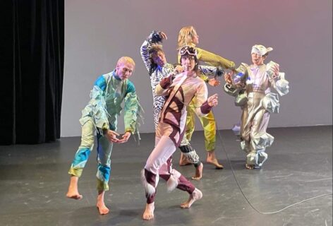 A performance photo of 5 dancers in various funky colored fabrics and patterns of animal like costumes; a performer in the center holds a microphone and wears a pink colored suit with matching goggles. Photo courtesy of the Artist.