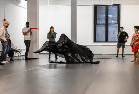 From the MR Archives 2022, Open Performance: A performer draped in black sheer fabric in motion in the center of the studio floor, an audience observes on the perimeter. Photo by Rachel Keane.