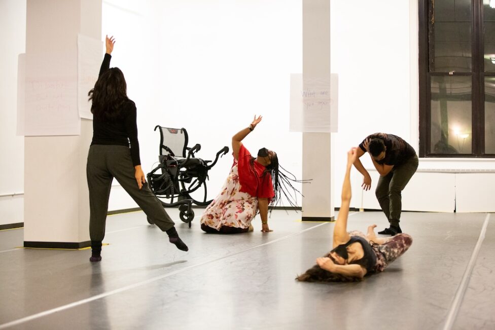 Photo from Elisabeth Motley and Kayla Hamilton’s AMP Residency showing. Four dancers move around the space, two closer to the floor and the other two on their feet. A wheelchair is parks in the background. Photo by Whitney Browne.