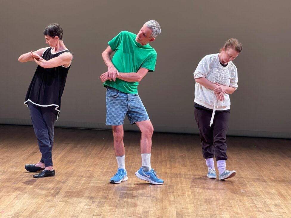 Three figures pose mid-movement atop a wooden floor, each grasping their respective wrists with opposite hands, heads tilted this way and that. Photo credit: Deborah Hay