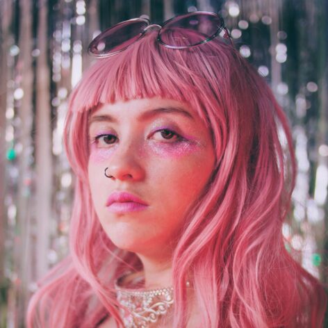The image features an artist's portrait with a retro and androgynous aesthetic, wearing a pink wig, glittery makeup, and a velvet outfit in shades of pink. The shimmering, blurred background, along with accessories like glasses and an ornate necklace, enhance a dreamy and nostalgic atmosphere. Photo taken by Luisa Ramírez.