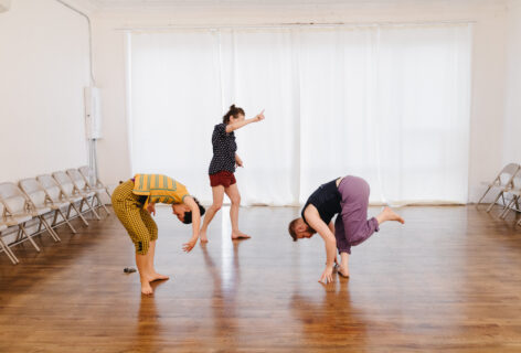 Three dancers move upon an shiny wooden floor. Two of them curl over and into themselves, and one stands with right arm outstretched, index finger pointing up. Photo credit to Lory Lyon.