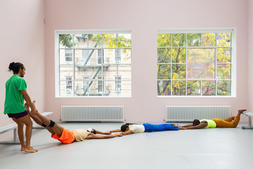 A room with pale pink walls and large windows frames four brightly costumed dancers. Three of the dancers lay horizontally across the diagonal of the floor, holding one another's limbs, while one dancer stands and holds the ankles of the last person in the chain. Photo credit to Helena Goñi. Ligia Lewis' study now steady at CARA, NYC.