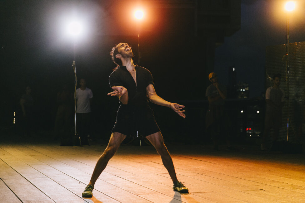 Miguel stands bathed in orange and yellow light, feet planted in a wide stance, head and hands offered skyward. Their eyes are closed and their mouth is open as if crying out or speaking. Photo credit to Rowa Lee. Ligia Lewis' Sensation 1 / This Interior at The High Line NYC. 