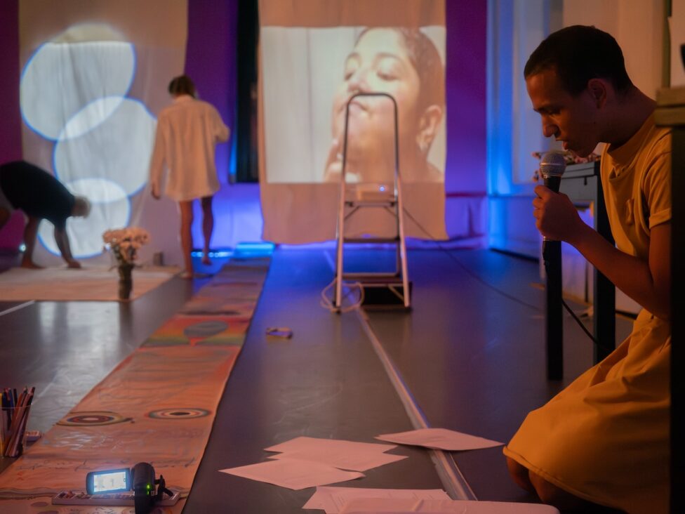 Photo from Buffy, stefa, and Paris’ AMP Residency showing: A studio is filled with lights, materials and projections in the background, while Paris kneels and talks into a mic and Buffy and stefa move in the background.