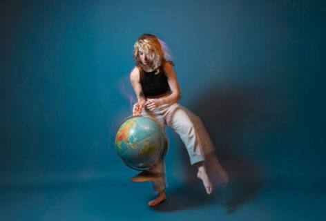 Rachel is dancing in front of a bright blue background with a globe. Their face and body is captured sharply, but they leave a trail of motion behind them. Self Portrait by Rachel Keane.