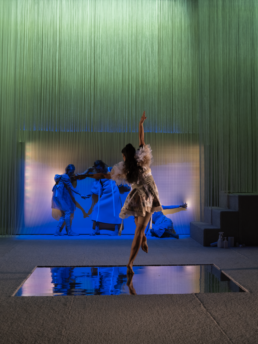A dancer (devika) extends their right arm to the sky while perched on the ball of their right foot. Below them is a square of a reflective surface, and they face a soft green, lavender, and yellow backdrop. Photo credit to Kameron Herndon. Big Dance Theater, 