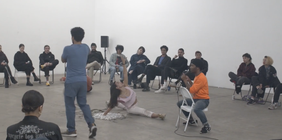 Wearing a pale purple shirt and white pants, devika poses on the ground in a split, their left arm reaching upwards. Along the perimeter of a large white wall, people sit in chairs and watch. Photo credit to Tom Tsai. Ajani Brannum, 