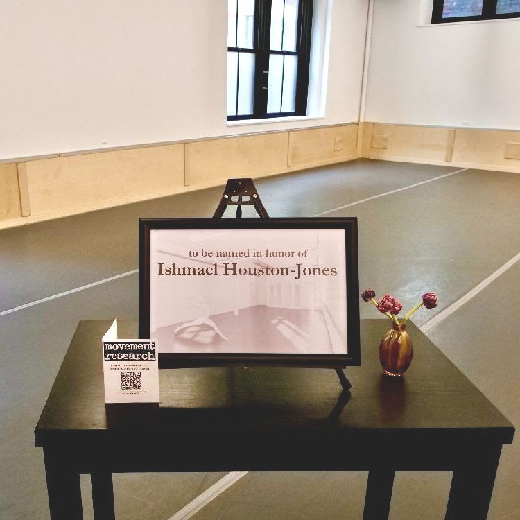 In a dance studio with a grey marley floor and one window in the back left corner there is a black desk. On the desk is (from left to right) is a Movement Research pamphlet with a QR code, a picture frame holding a black and white image of a dance studio with the words “to be named in honor of Ishmael Houston-Jones” in black text, and a purple glass vase with purple flowers.