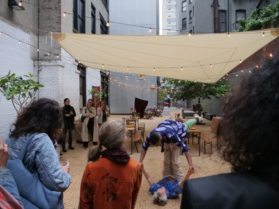 Photo from the 2024 MR Groundbreaking Fundraiser; People stand in the 122CC Courtyard looking toward two performers, one performer balances on the legs of the second performer who lies on the ground. Photo by Katherine De La Cruz