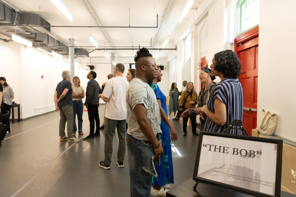 In a dance studio with red doors and a grey marley floor 12 middle aged people of mixed races stand mingling with smiling and expressive faces. There is a picture frame in the bottom right hand corner on a black desk. In the frame is the image of a dance studio in black and white with “THE BOB” written in capital letters and black text. Photos by Daniela Garza Maldonado.