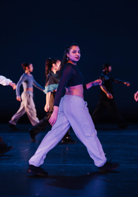 Rose wears a dark blue mesh crop top, paired with low waisted light blue pants and black sneakers. She is strutting across the stage with her head turned towards the audience. Other dancers are blurred in the background while the camera is focused on her. Photo credit: Chris Kayden