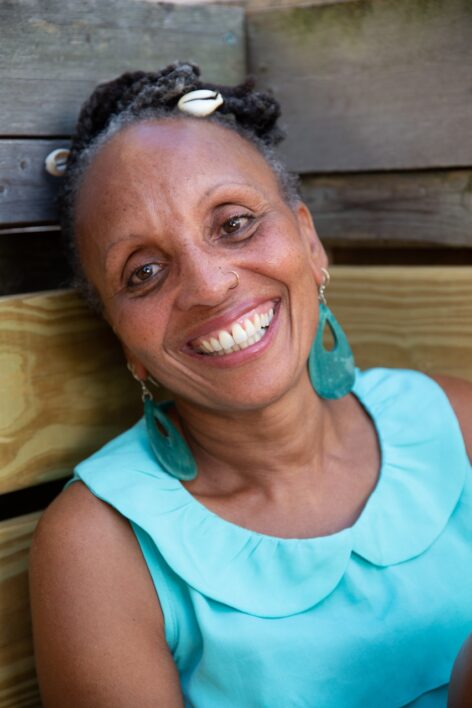Charmaine Warren in a bright blur top smiles at the camera in front of a slated wall, she wears teal earrings and her hair is up adorn with shells. Photo by Tony Turner.