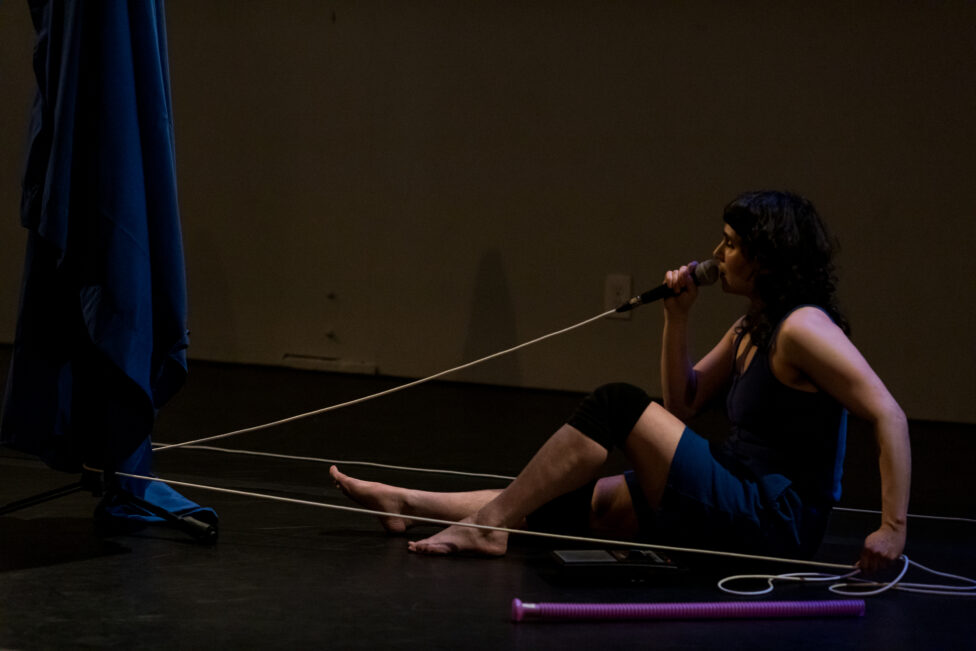 Sitting with her right leg extended, Allie holds a microphone up to her mouth. A chord extends from the microphone and disappears behind a curtain. She pulls the rest of the chord taught with her left hand. 