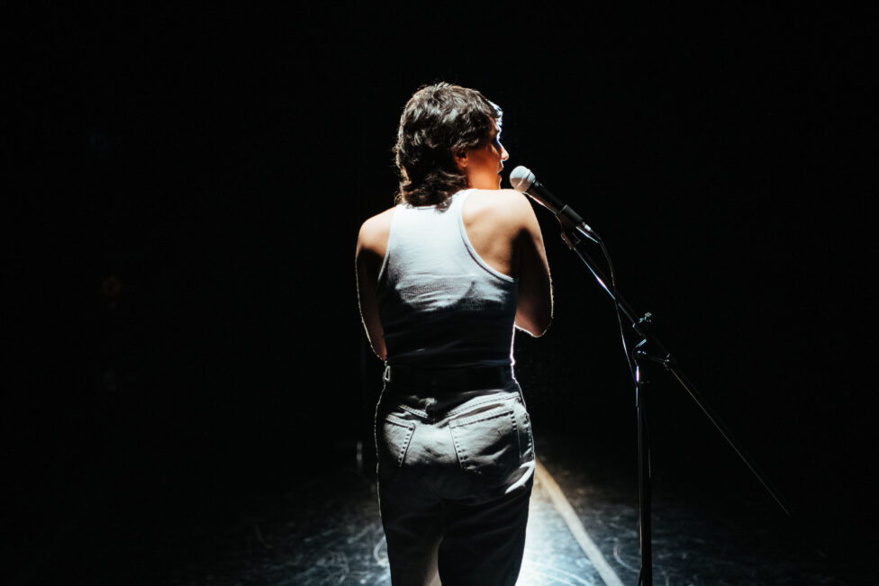 Allie stands facing away from the camera with her head turned to the right, revealing one side of her face. She wears a white tank top and jeans and is shrouded in shadow and light. The background is an inky black. 