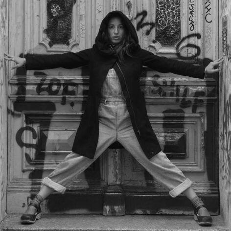 A black-and-white photo of Verónica. A woman standing with her arms open, fingertips touching the walls on either side. Her legs are spread wider than shoulder-width apart, in front of a closed door covered in graffiti. She faces forward, creating a sense of expansion and presence. She wears denim overalls and a winter jacket with a hood that covers her head, though her hair remains visible, in contact with her heart. Photo by Alexander Carrillo.