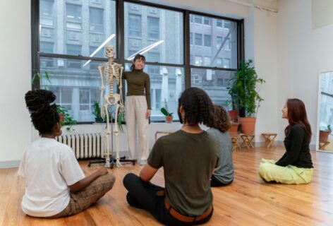 Holly is smiling and touching the spine a life-sized human skeleton as she faces a class full of students who are seated on the floor. Photo by Bryan Bisordi.