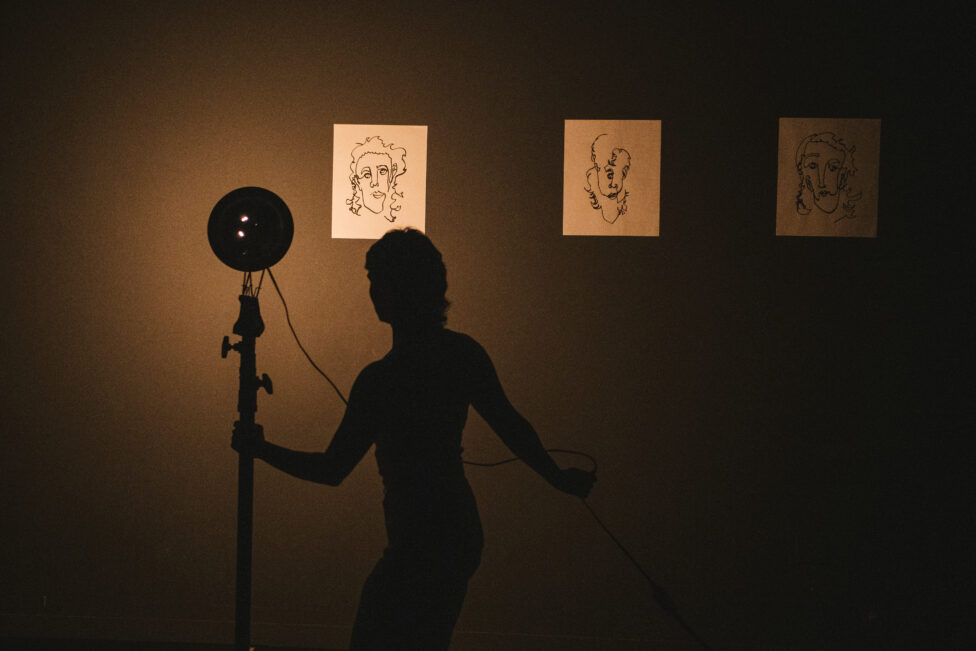 A silhouetted figure poses against a warmly lit backdrop. Behind her are drawings of faces on white paper. 