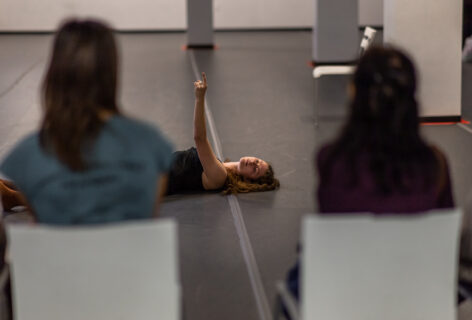 A photo of an Open Performance artist reaching their arm up and laying on the ground. In the foreground the audience turns its back to the camera viewing the performance. Photo by Rachel Keane.