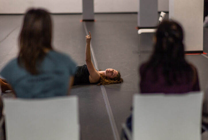 A photo of an Open Performance artist reaching their arm up and laying on the ground. In the foreground the audience turns its back to the camera viewing the performance. Photo by Rachel Keane.