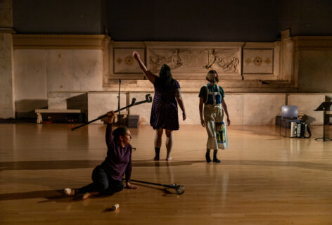 Kiera Bono, Michelle Mantione and Krista Miranda perfoming for Movement Research at the Judson Church. One dancer sits on the ground holding one crutch above their head and resting the other on the ground. Two other artists stand nearby. One waves their arm upwards. Photo by Rachel Keane.