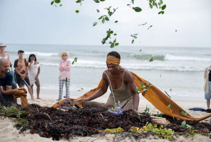 black artist kneeling in the sand on a beach with an elongated golden blindfold surrounded by a small group of people. Ocean in the background. Artist sitting in front of a mound of seaweed and plant parts captured in a gesture of throwing, leaves suspended overhead. Dynamic colors of ocean blue, green leaves, brown sand, the artist brown skin and the silver shimmer of the sun on the water in the background. Photo by Nir Arieli.