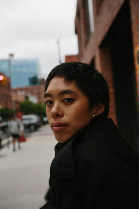 Michaela, a Filipina woman with short black hair, looks back at the camera. She is wearing a black jacket with a high neckline and is not smiling. 