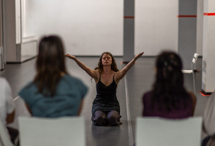 An Open Performance artist sits with their arms stretched out and upward. In the foreground the audience turns its back to the camera viewing the performance. Photo by Rachel Keane