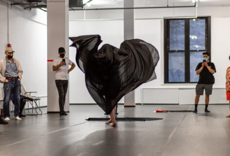 A photo of an Open performance artist: The artist stands in the middle of the apace and a sheer black fabric draped over their body billows out all around them. Photo by Rachel Keane.