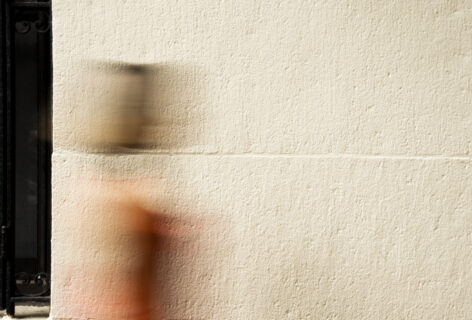 The photo shows a blurred figure in motion walking past a textured, beige wall. The person is wearing a red outfit, with their head and body smeared across the image as a result of a slow shutter speed. On the left side of the image, there is a black window with vertical iron bars, contrasting against the smooth wall and the movement of the figure. Photo By Rojin Shafiei.